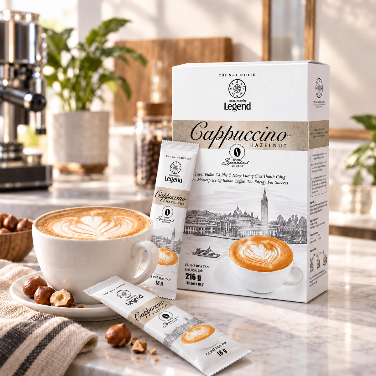 A box and two sachets of Legend Cappuccino Hazelnut instant coffee are displayed on a marble countertop next to a cup of latte with heart-shaped foam art, with hazelnuts and a coffee machine blurred in the background.
